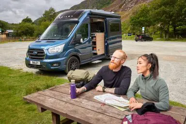 A couple sat on a picnic bench at a rest stop outside their Bailey Endeavour campervan