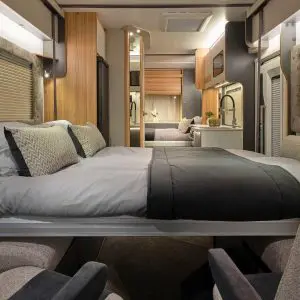 Interior view of a motorhome bedroom – A modern and stylish motorhome bedroom featuring a double bed with grey and black bedding, plush pillows, and warm ambient lighting. The design incorporates wood paneling and mirrored surfaces, enhancing the sense of space.