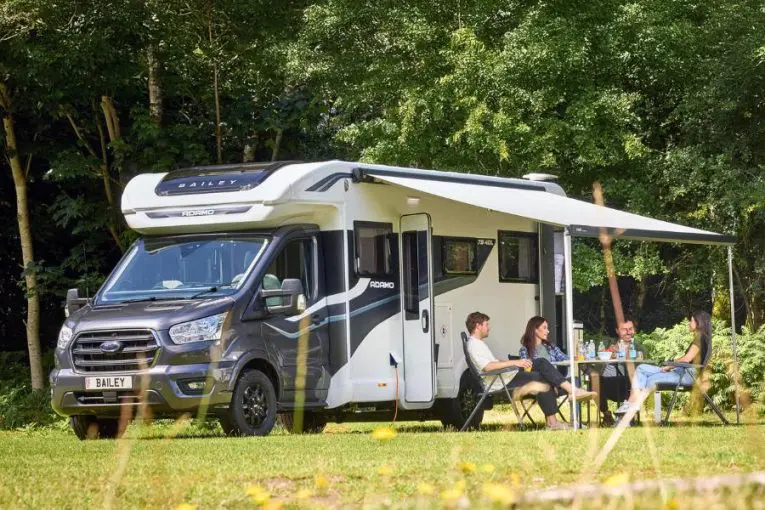 Outdoor camping scene with a Bailey Adamo motorhome – A group of four people relaxing under the extended awning of a Bailey Adamo motorhome, enjoying an outdoor meal in a lush, wooded area. The vehicle is fully equipped for comfortable travel and leisure.