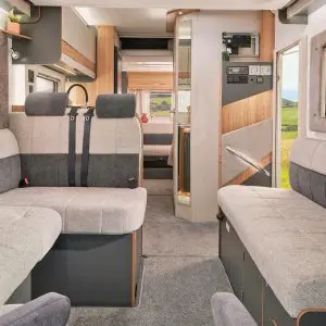 Luxury lounge area in a motorhome – A well-lit and spacious seating area in a motorhome, featuring plush, dual-tone upholstery, large windows, and a view extending towards the kitchen and sleeping area.