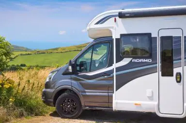 Side view of an Adamo motorhome in a scenic location – A sleek, modern Bailey Adamo motorhome parked in a countryside setting with rolling green hills and the ocean in the distance. The black and white exterior design features stylish branding.