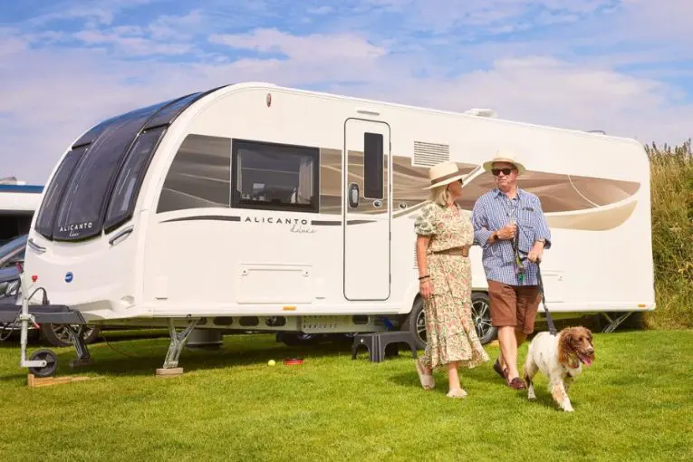 Elderly couple walking their dog beside a Bailey Alicanto Grande Deluxe caravan pitched on grass, with sunny weather and blue skies.