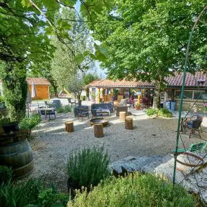 An inviting outdoor communal area at Domaine de la Faurie featuring rustic wooden seating, potted plants, and several stone and timber buildings shaded by leafy trees.