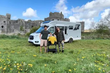 A young couple posing with a baby stroller in front of a Bailey Alora motorhome, parked in a grassy field near a historic castle ruin.