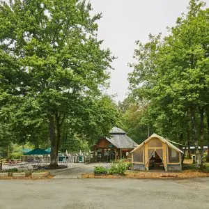 Central communal facilities at Huttopia Lac de Sillé, including a wooden lodge, canvas tent, and outdoor seating area, surrounded by tall green trees.
