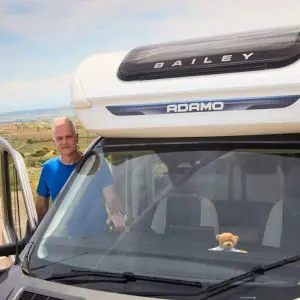 A man in a blue t-shirt stands beside an open door of a Bailey Adamo motorhome parked in a scenic rural location. A small teddy bear is visible on the dashboard inside the vehicle.