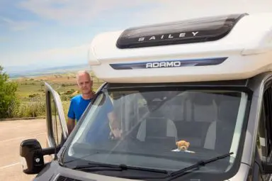 A man in a blue t-shirt stands beside an open door of a Bailey Adamo motorhome parked in a scenic rural location. A small teddy bear is visible on the dashboard inside the vehicle.