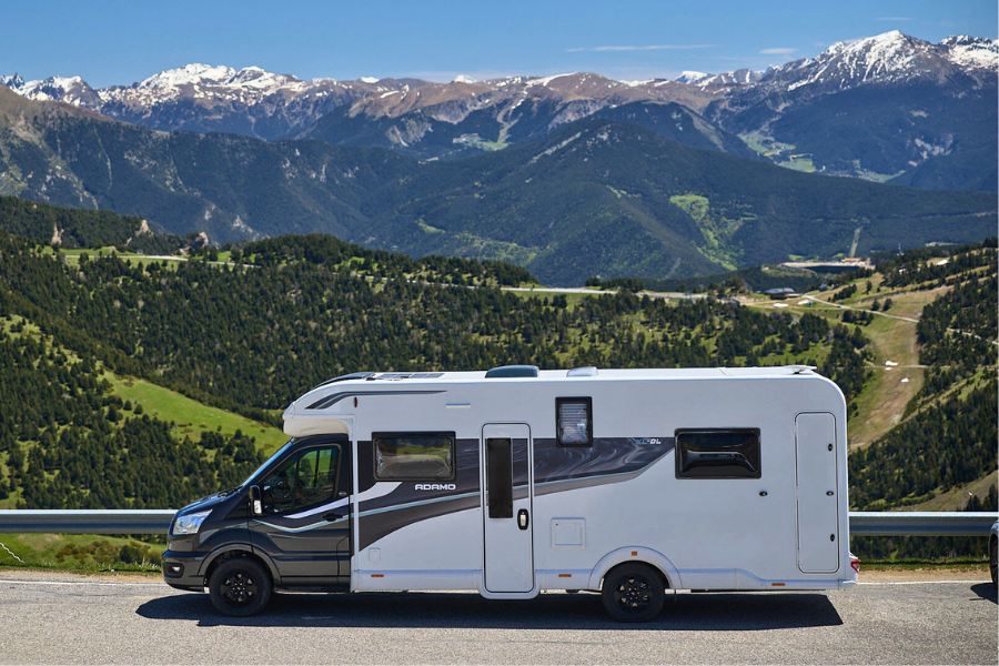 Bailey Adamo XL-DL motorhome driving along a mountain road with scenic views of alpine forests and snow-capped peaks in the background.