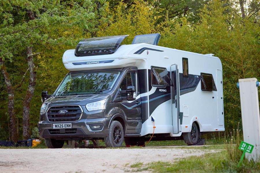 Bailey Adamo XL-DL motorhome parked on a campsite surrounded by trees, with electric hookup connected and roof lights open.