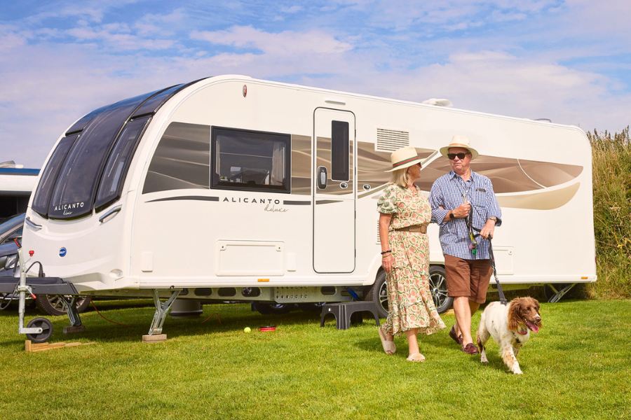 Elderly couple walking their dog beside a Bailey Alicanto Grande Deluxe caravan pitched on grass, with sunny weather and blue skies.