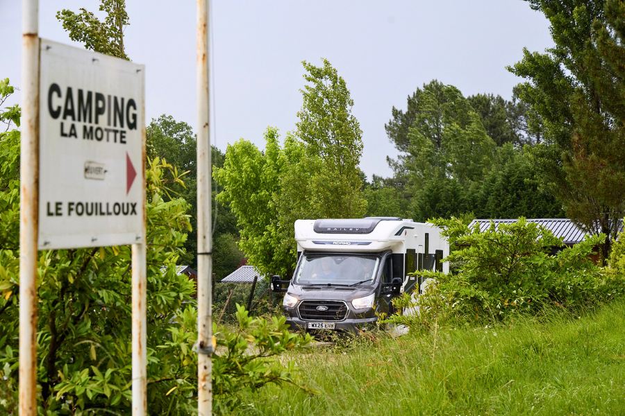 A Bailey Adamo motorhome drives through a lush green entrance to Camping La Motte in Le Fouilloux, France, with a white entrance sign visible in the foreground.