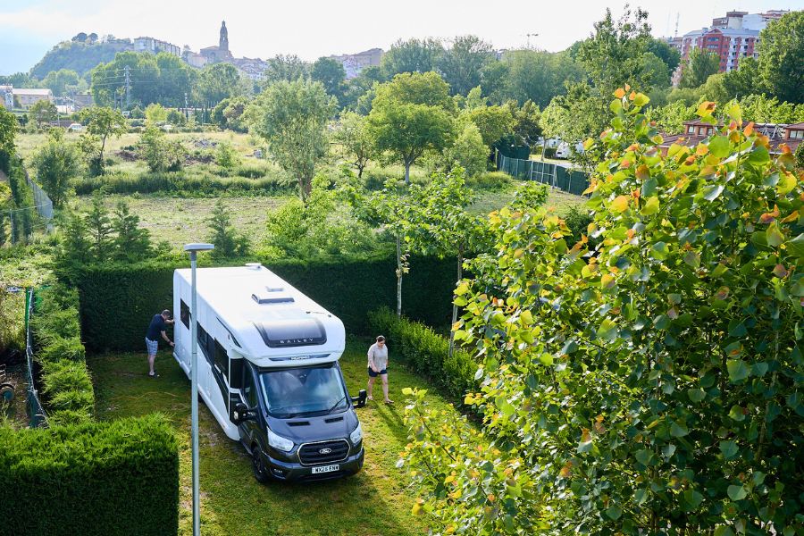 A Bailey motorhome is parked on a neatly trimmed pitch at Camping De Haro, surrounded by hedges and greenery, with two people setting up nearby under a bright sky.