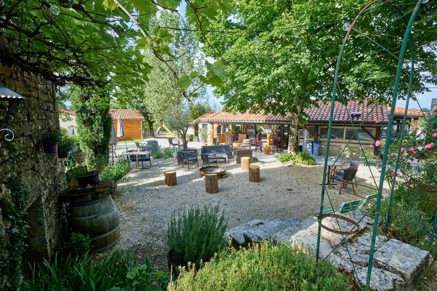 An inviting outdoor communal area at Domaine de la Faurie featuring rustic wooden seating, potted plants, and several stone and timber buildings shaded by leafy trees.