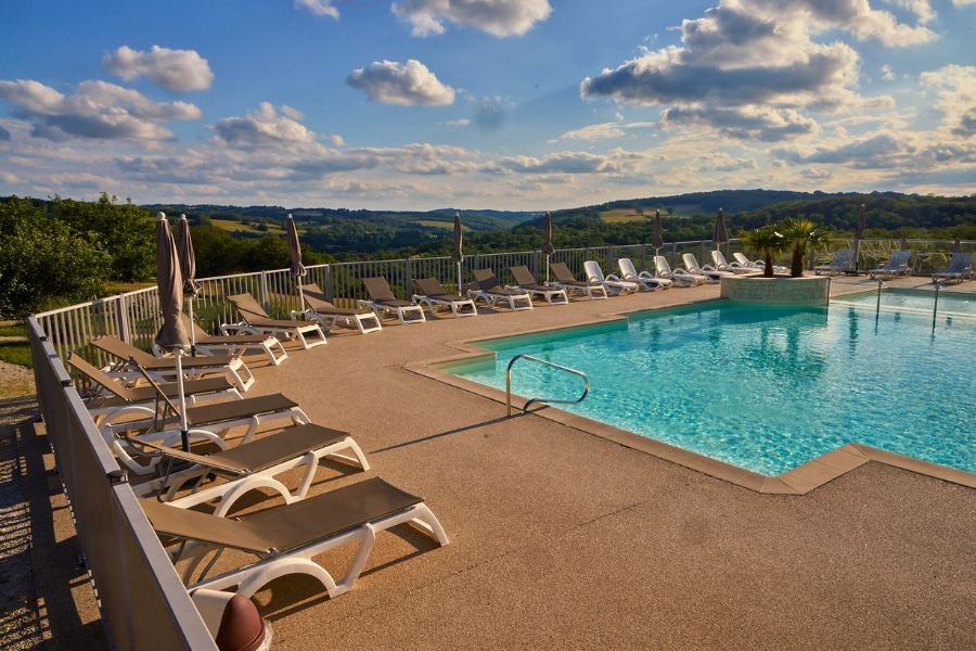 A serene swimming pool at Domaine de la Faurie with rows of sun loungers and parasols, overlooking scenic countryside hills under a partly cloudy sky.