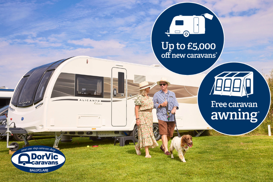 A couple walks their dog next to a Bailey Alicanto Grande caravan on a sunny day. Promotional graphics highlight “Up to £5,000 off new caravans” and “Free caravan awning.” The Dorvic Caravans Ballyclare logo is also visible.
