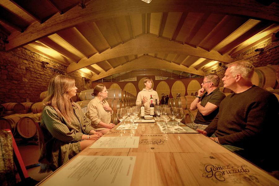 Wine tasting session at Gómez Cruzado winery, with a guide presenting to five visitors seated around a table in a warmly lit cellar surrounded by wooden barrels.
