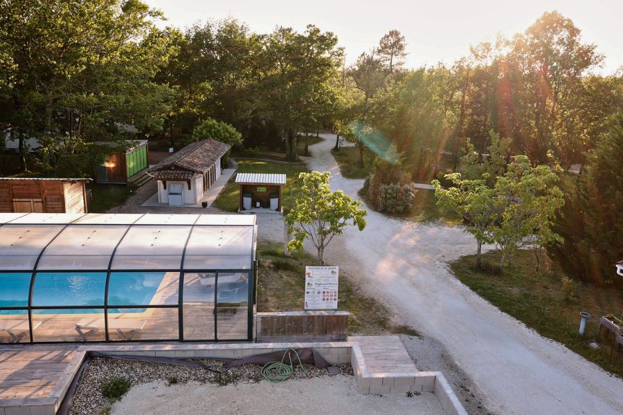 An aerial view of Camping La Motte showing a covered swimming pool, rustic buildings, and gravel paths winding through trees in warm evening sunlight.