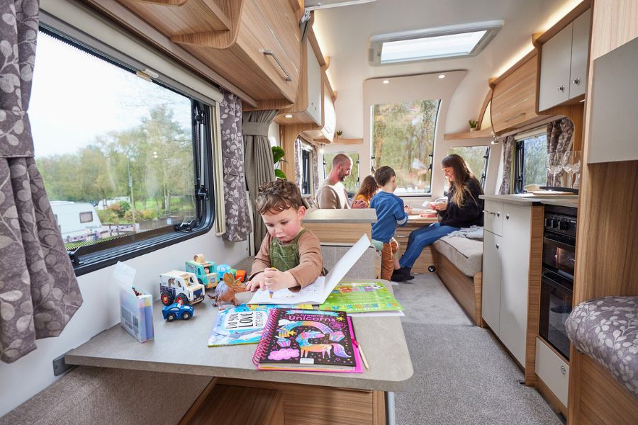 Family with young children enjoying time inside a Bailey Phoenix GT75 caravan, with a child drawing at a table and parents dining in the background.