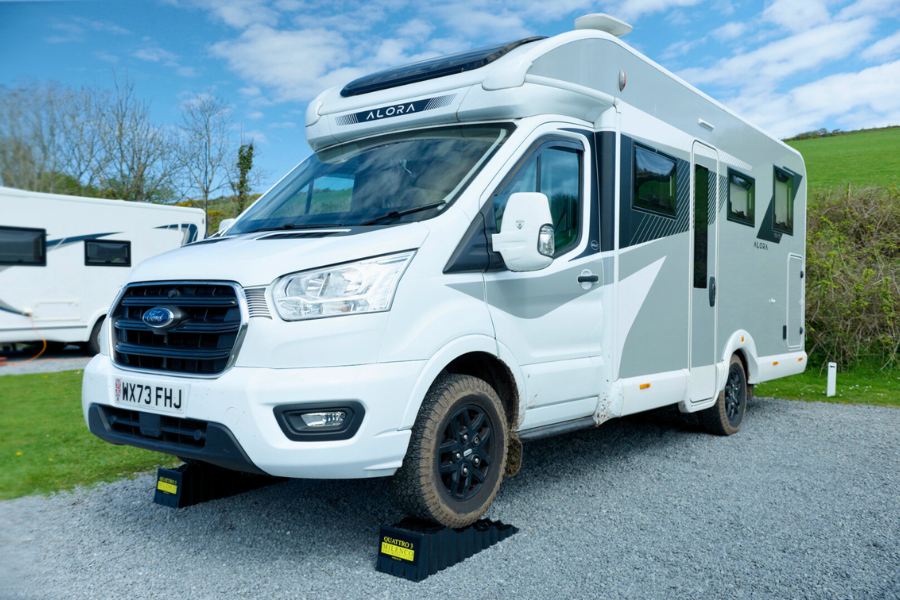A Bailey Alora motorhome parked on a gravel pitch with its wheels on levelling ramps, surrounded by green countryside under a blue sky.
