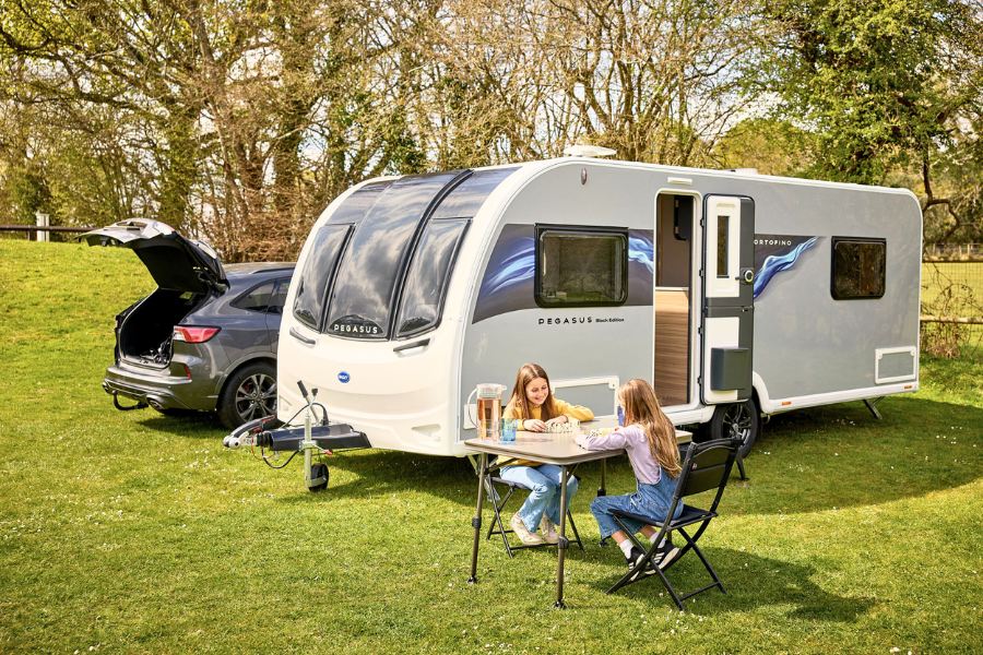 The Pegasus Black Edition Portofino caravan pitched on grass with a car. Two children sit at a table enjoying the outdoors, highlighting its family-friendly design.